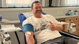 Ein Mann in einem Krankenhausstuhl, der ein weißes T-Shirt mit einem grafischen Design trägt, lächelt und gibt einen Daumen hoch. Er hat eine Blutdruckmanschette am linken Arm.
