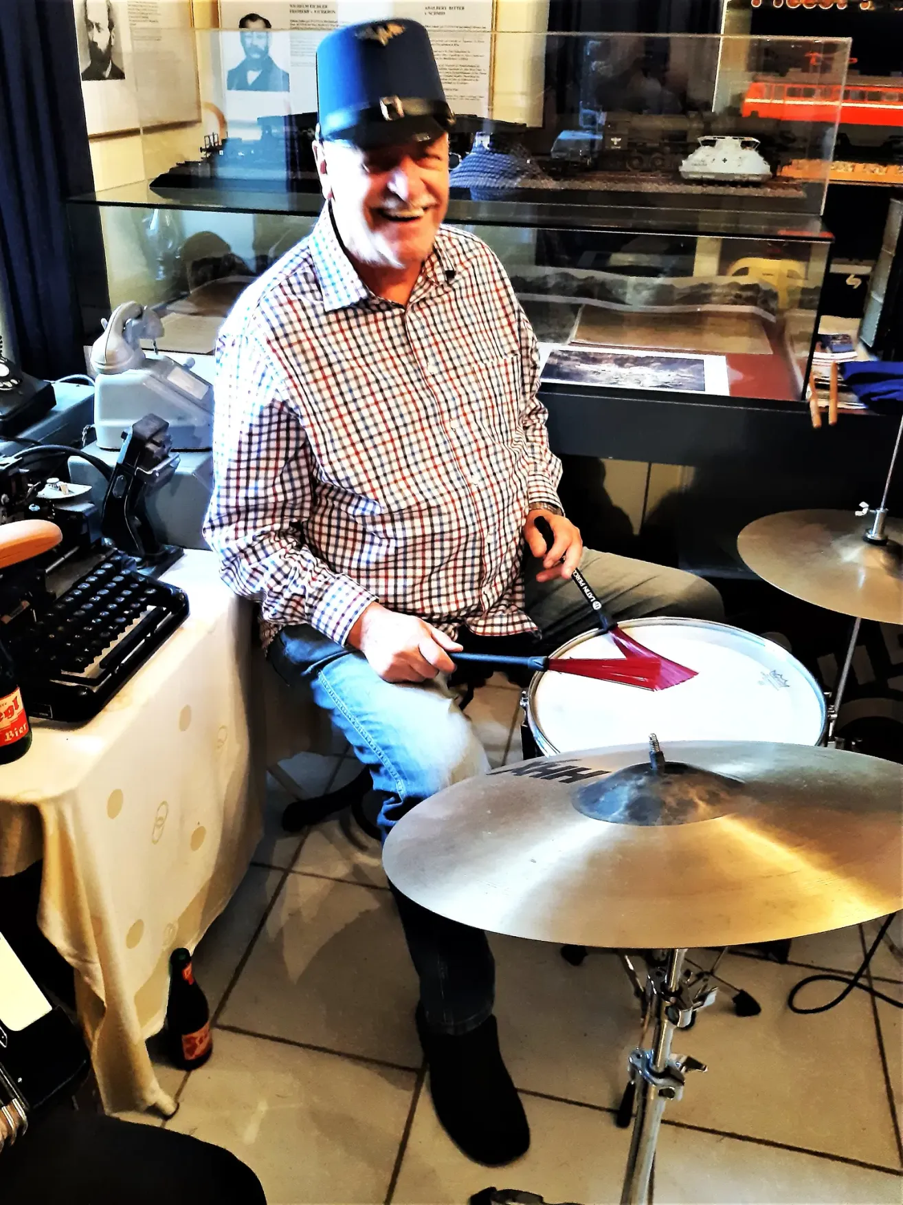 Ein Mann sitzt auf einem Hocker, lächelt und spielt eine Trommel mit einem Drumstick in der Hand. Hinter ihm steht eine Glasvitrine mit verschiedenen Gegenständen, einer Schreibmaschine und einer Flasche auf einem Tisch.
