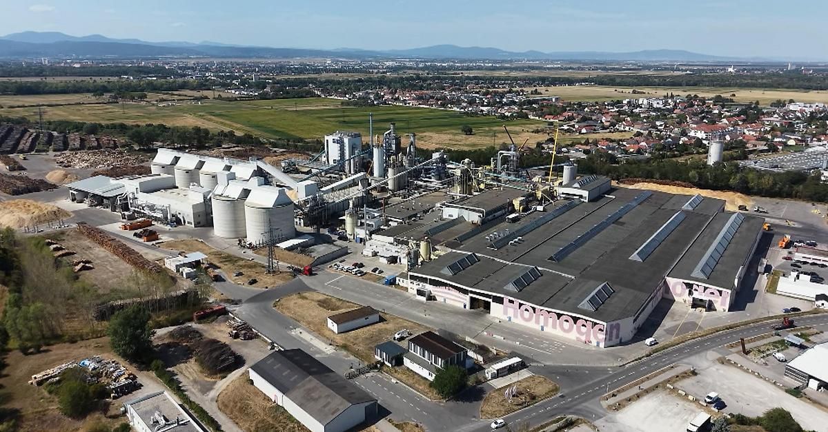Luftaufnahme einer großen Industrieanlage mit mehreren Gebäuden, großen Silos und Solarpanels auf dem Dach. Die Anlage ist von einem weitläufigen grünen Feld und einer Stadt in der Ferne umgeben.