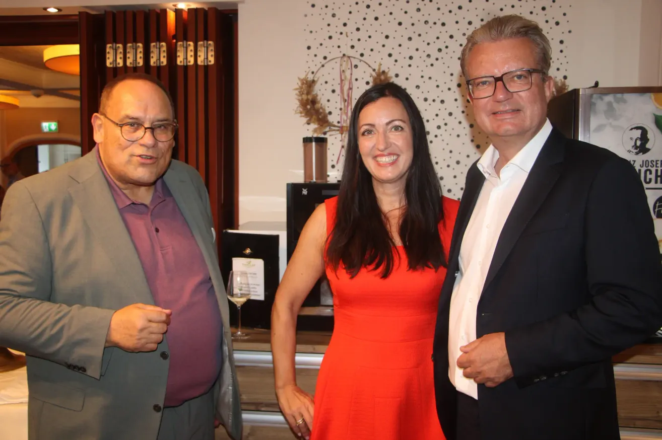 Three people stand close together, smiling for a photo. The woman in the middle wears a red dress. The man on the left wears a gray suit, and the man on the right wears a black suit. Behind them is a wall with black and white dots.