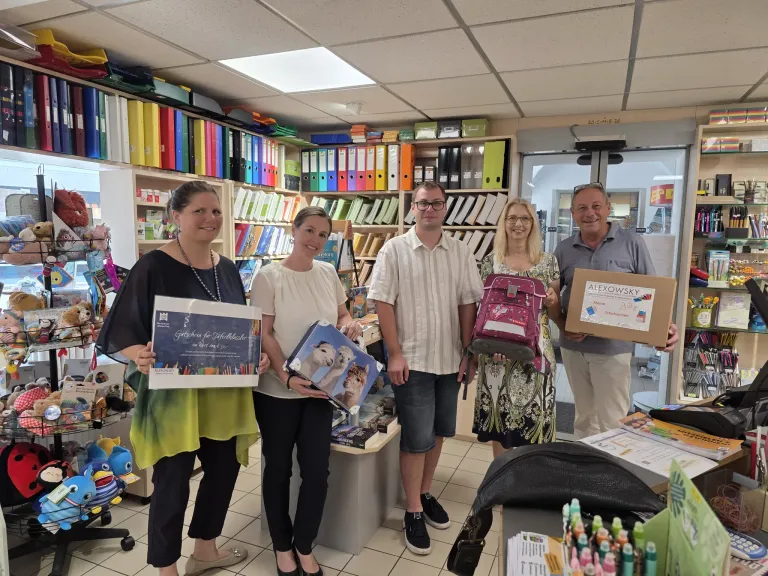 Five people are standing in a room with shelves of books and folders in the background. They are holding various items, including a certificate, a box, and a backpack. The woman on the left is wearing a necklace and a green top. The man in the middle is wearing glasses and a white shirt. The woman on the right is wearing a floral dress and holding a backpack.