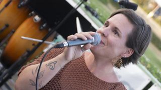 Eine Frau mit Tattoos am Arm singt in ein Mikrofon, während sie ein braunes Oberteil und goldene Ohrringe trägt.