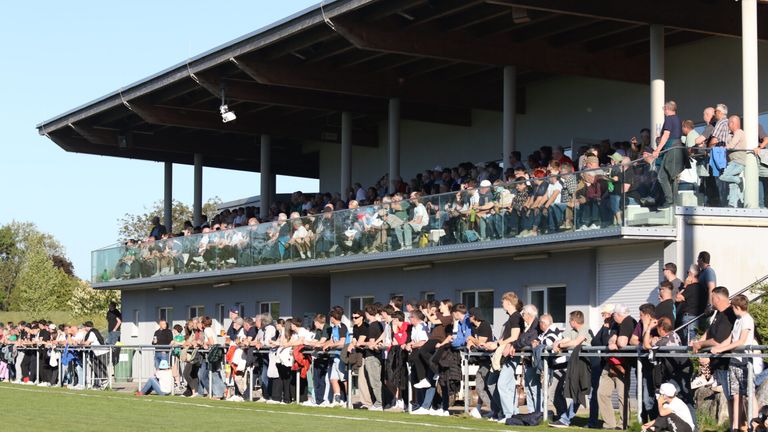 A crowd of people watching a game from the stands and on the field, with some holding jackets and bags on the railings.