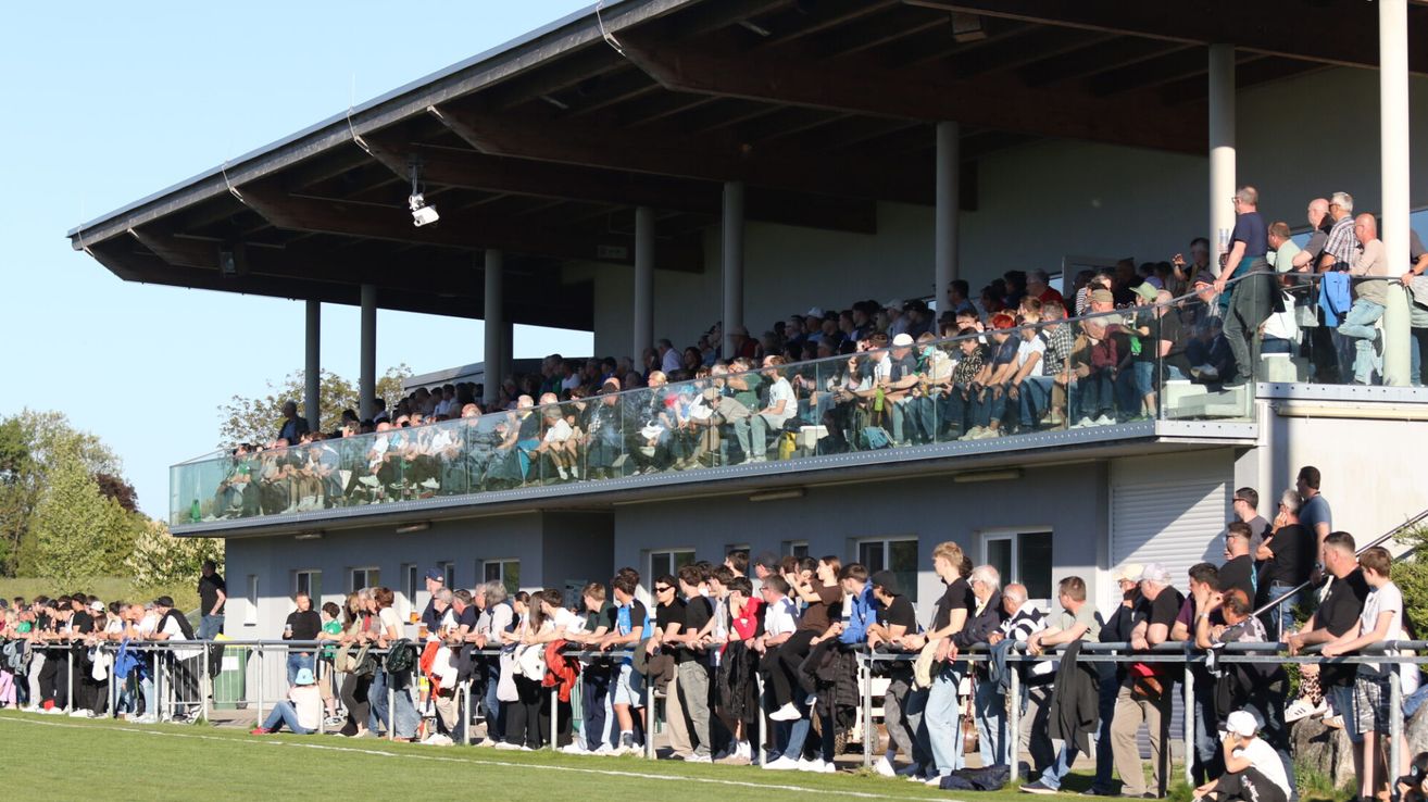 A crowd of people watching a game from the stands and on the field, with some holding jackets and bags on the railings.