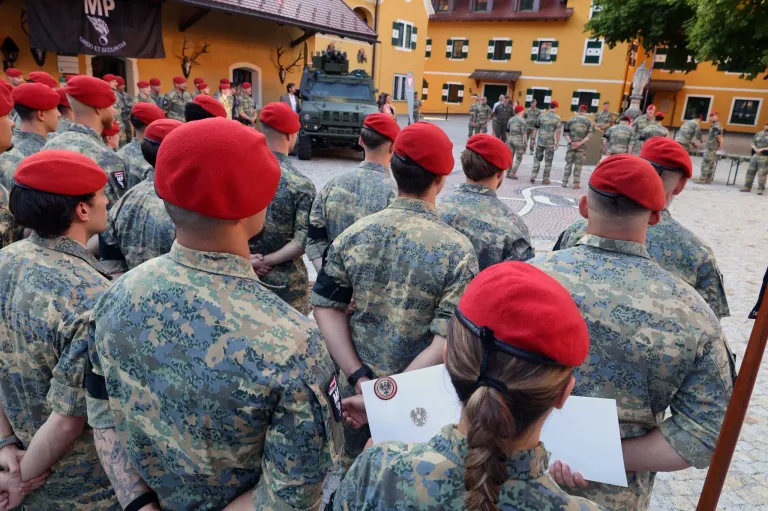 Eine Gruppe von Militärangehörigen in Tarnuniformen und roten Baretten steht in Formation, mit einem Militärfahrzeug und gelben Gebäuden im Hintergrund.