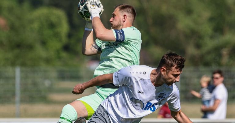 A soccer player in a green jersey is catching a ball while another player in a white jersey tries to steal it.