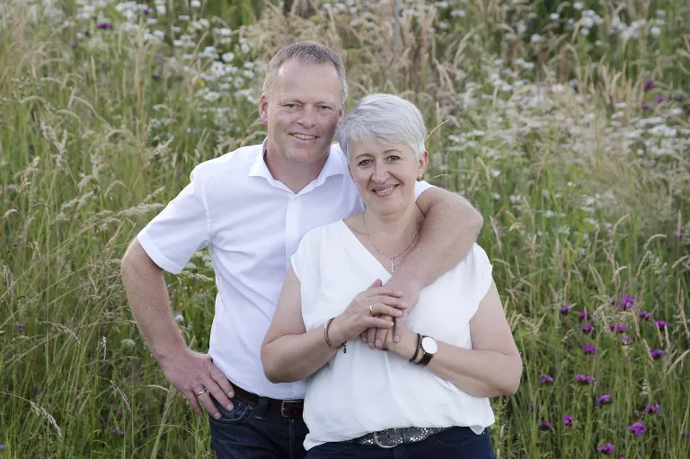 Ein Mann und eine Frau stehen in einem Feld mit hohem Gras und posieren für ein Foto. Der Mann hat seinen Arm um die Schulter der Frau gelegt.
