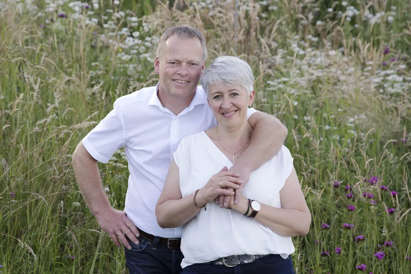 Ein Mann und eine Frau stehen in einem Feld mit hohem Gras und posieren für ein Foto. Der Mann hat seinen Arm um die Schulter der Frau gelegt.