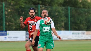Zwei Fußballspieler in Aktion auf einem Feld, einer in Rot und der andere in Grün, wobei der Spieler in Grün kurz davor steht, den Ball zu schießen.