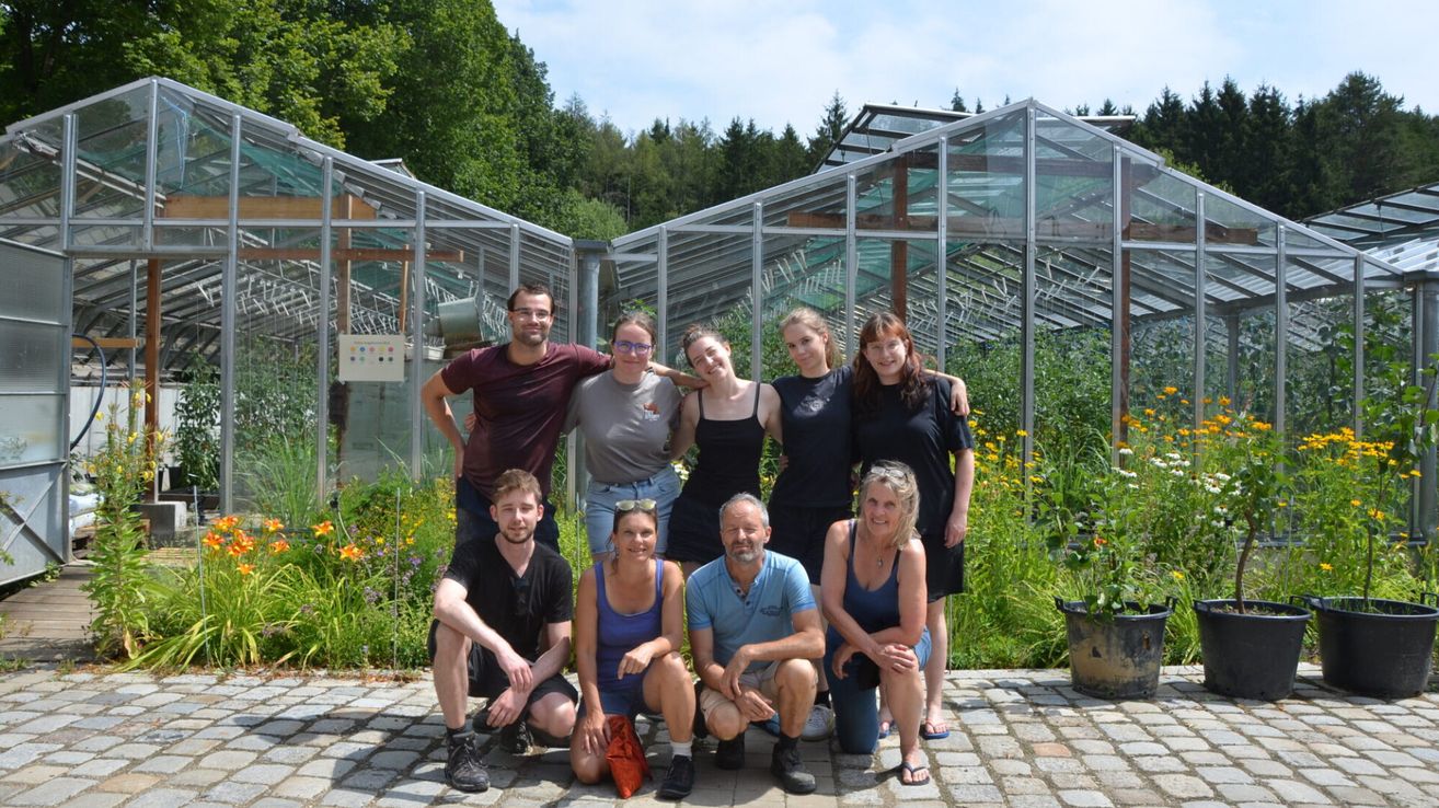 A group of people are posing for a photo in front of a greenhouse with glass walls and a metal frame. They are standing and sitting on a brick floor, with some wearing glasses and sneakers. Behind them are plants and trees.