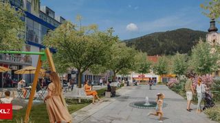 Ein belebter Stadtplatz mit Menschen, die gehen, sitzen und ein Kind, das auf einen Brunnen zuläuft. Bäume und Gebäude säumen die Straße, mit Bergen im Hintergrund.