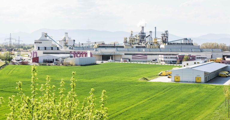 Ein großes Industriegelände mit mehreren Gebäuden und Maschinen, umgeben von einem weitläufigen grünen Feld unter einem bewölkten Himmel.