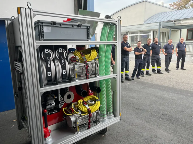 A group of firefighters stand next to a wheeled cart filled with equipment, including hoses and cylinders labeled 'B'.