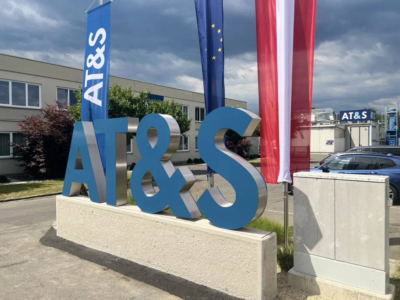 Ein großes metallisches Schild für AT&S steht vor einem Gebäude mit Fahnen dahinter. Das Schild ist blau mit weißen Buchstaben und steht auf einem Betonsockel. Dahinter befinden sich zwei Fahnen, eine davon ist die EU-Fahne.