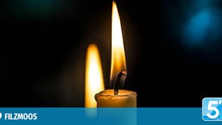 Eine Nahaufnahme einer brennenden Kerze mit einer leicht gebogenen Flamme. Die Kerze ist gelb und hat eine glatte Oberfläche. Der Hintergrund ist dunkel.