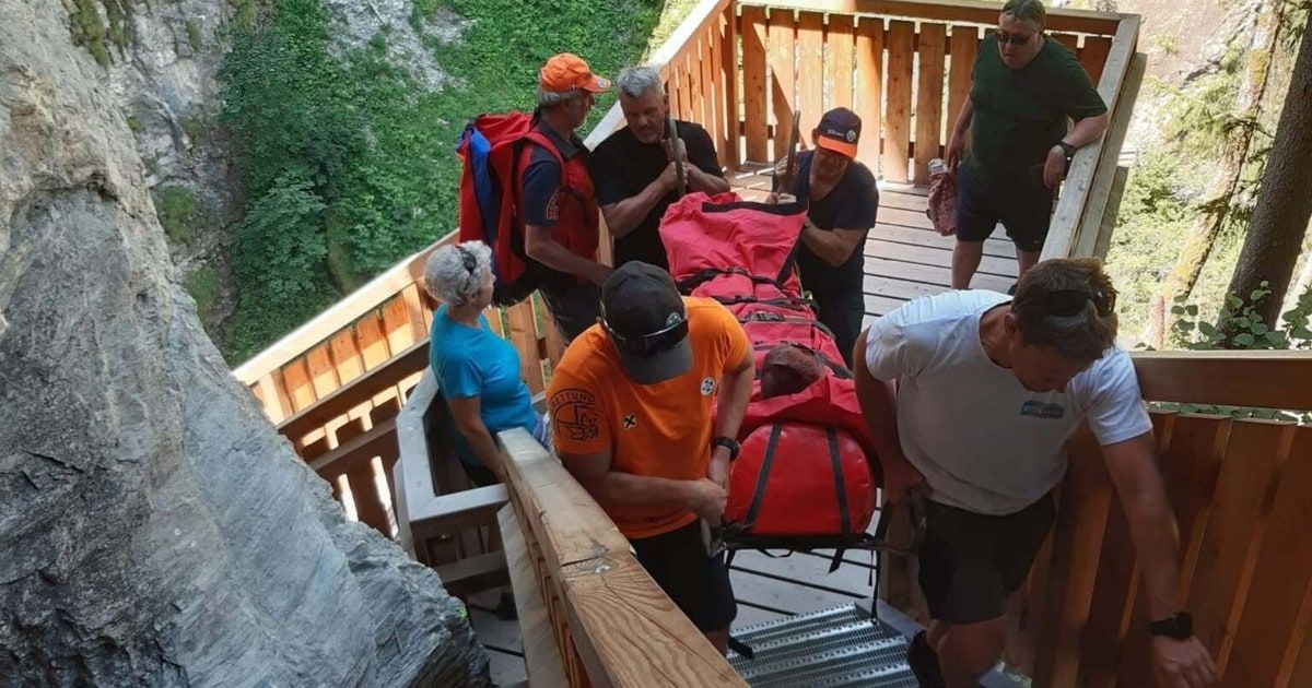 Eine Gruppe von Menschen in orangefarbenen und schwarzen T-Shirts trägt eine rote Trage eine Holztreppe hinunter.