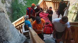 Eine Gruppe von Menschen in orangefarbenen und schwarzen T-Shirts trägt eine rote Trage eine Holztreppe hinunter.