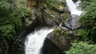 Ein Wasserfall stürzt an einem felsigen Abhang in einem üppigen, grünen Wald hinab, wobei das Wasser auf die Felsen darunter spritzt.