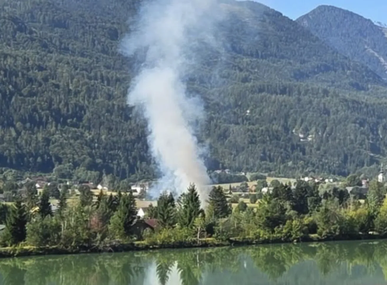 Ein großer Rauchschwaden steigt von einem bewaldeten Hang auf, in der Nähe eines Flusses und einer kleinen Stadt.