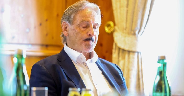 Ein älterer Mann mit grauem Haar und einem Schnurrbart sitzt in einem Stuhl, trägt einen blauen Anzug und ein weißes Hemd. Er schaut mit geschlossenen Augen nach unten. Auf dem Tisch vor ihm stehen zwei Gläser mit gelber Flüssigkeit.