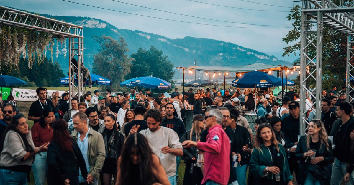 A large crowd of people are gathered outdoors, likely at a festival, with mountains in the background. They are under blue umbrellas and near food stalls.