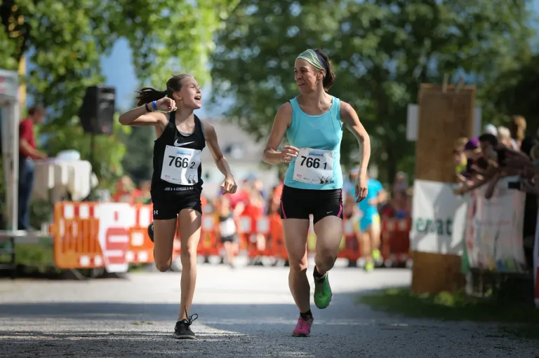 Zwei Frauen, eine mit der Nummer 7661 und die andere mit 7662, laufen bei einem Marathon, mit einem verschwommenen Hintergrund von Zuschauern und Bäumen.