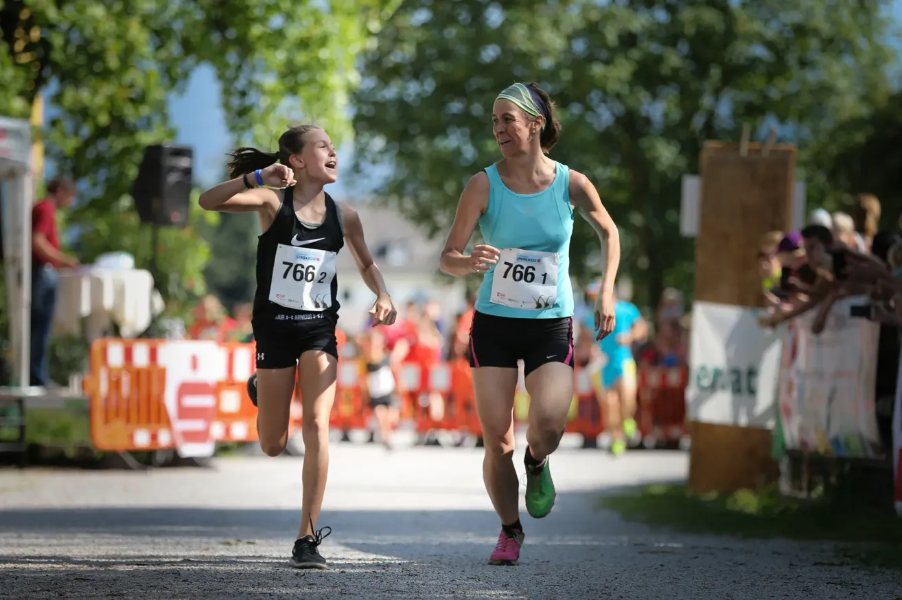 Zwei Frauen, eine mit der Nummer 7661 und die andere mit 7662, laufen bei einem Marathon, mit einem verschwommenen Hintergrund von Zuschauern und Bäumen.