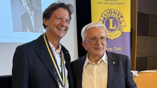 Zwei Männer stehen nebeneinander, lächelnd, vor einem Lions Club-Banner. Der Mann auf der linken Seite trägt eine Medaille.