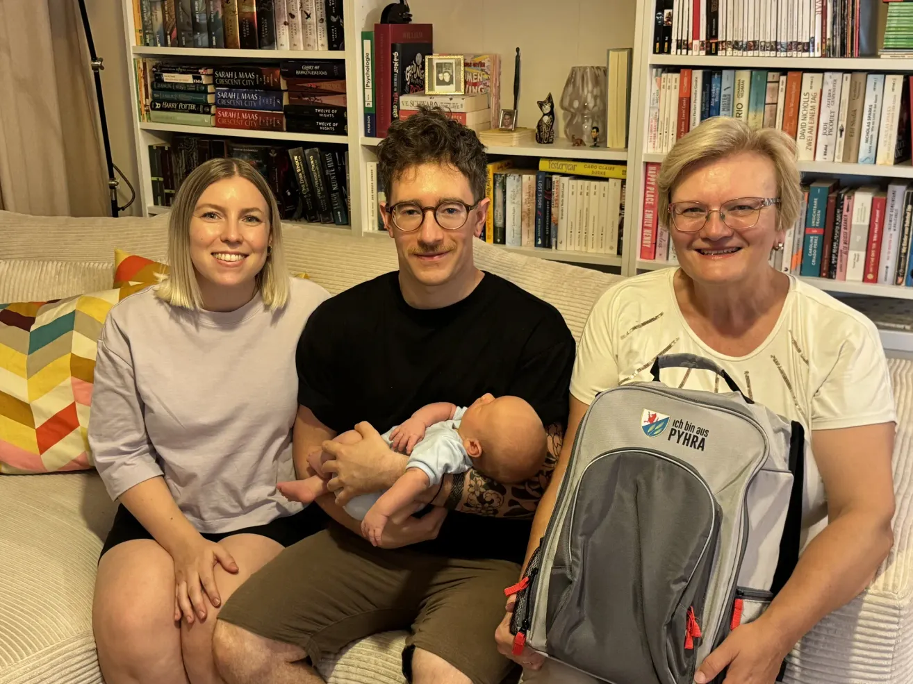Eine Familie aus drei Personen, darunter ein Mann, der ein Baby hält, eine Frau mit Brille und eine weitere Frau, die einen Rucksack hält, sitzen auf einem Sofa in einem Raum mit Bücherregalen voller Bücher.