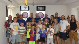 Eine Gruppe von Kindern und Erwachsenen posiert für ein Foto vor Dartbrettern und Monitoren und hält Pokale.