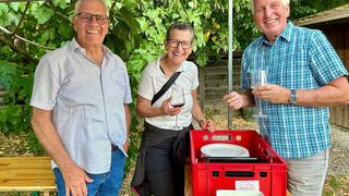 Drei Erwachsene lächeln und halten Weingläser in einer Gartenumgebung. Eine Frau in der Mitte hält ein Weinglas mit rotem Wein. Ein roter Kasten mit Tellern steht vor ihnen.