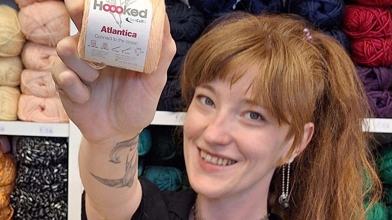 Eine Frau mit kurzen roten Haaren hält eine Spule Garn mit der Aufschrift 'Hooked Atlantica'. Sie lächelt und zeigt ein Tattoo auf ihrem Arm.