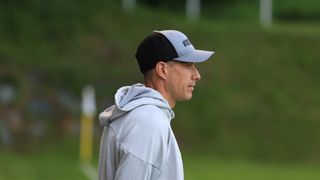 A man in a gray hoodie and a black and gray cap with the text '#SCH' is standing on a grassy field, possibly a sports field.