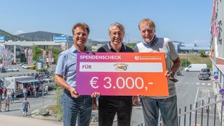 Drei Männer halten einen großen Scheck über 3.000 Euro und stehen auf einem Balkon mit einer malerischen Kulisse.