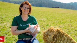 Eine Frau in einem grünen Shirt mit dem Wort 'Agarvision' hält ein kleines Schwein in einem grünen Feld mit einem Heuballen in der Nähe.