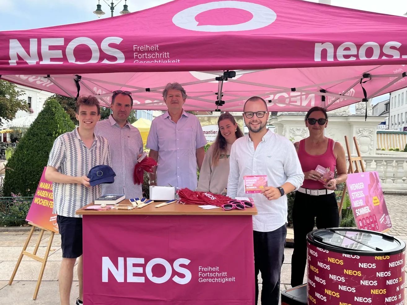 Eine Gruppe von Menschen steht unter einem rosa Zelt mit dem Wort NEOS darauf. Sie lächeln und posieren für ein Foto. Ein Tisch vor ihnen hat verschiedene Gegenstände darauf.