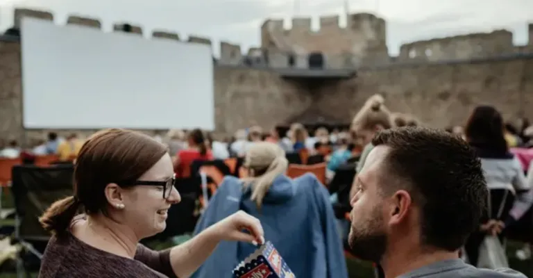 Eine Frau mit Brille hält eine Tüte Popcorn und lächelt einen Mann in einer Menschenmenge an, die einen Film im Freien ansieht.