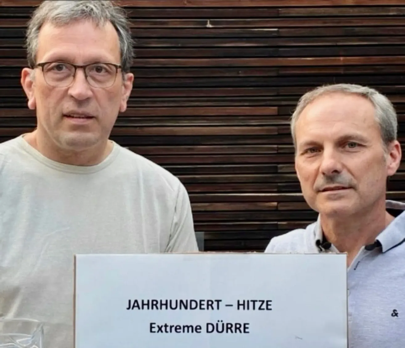 Two men stand next to each other with a sign that reads 'Jahrhundert Hitze Extreme Dürre' in front of a wooden wall.