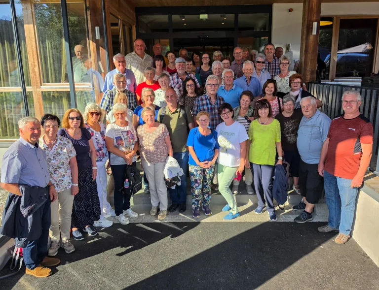 A group of people, mostly elderly, are standing in front of a building, smiling and posing for a photo.