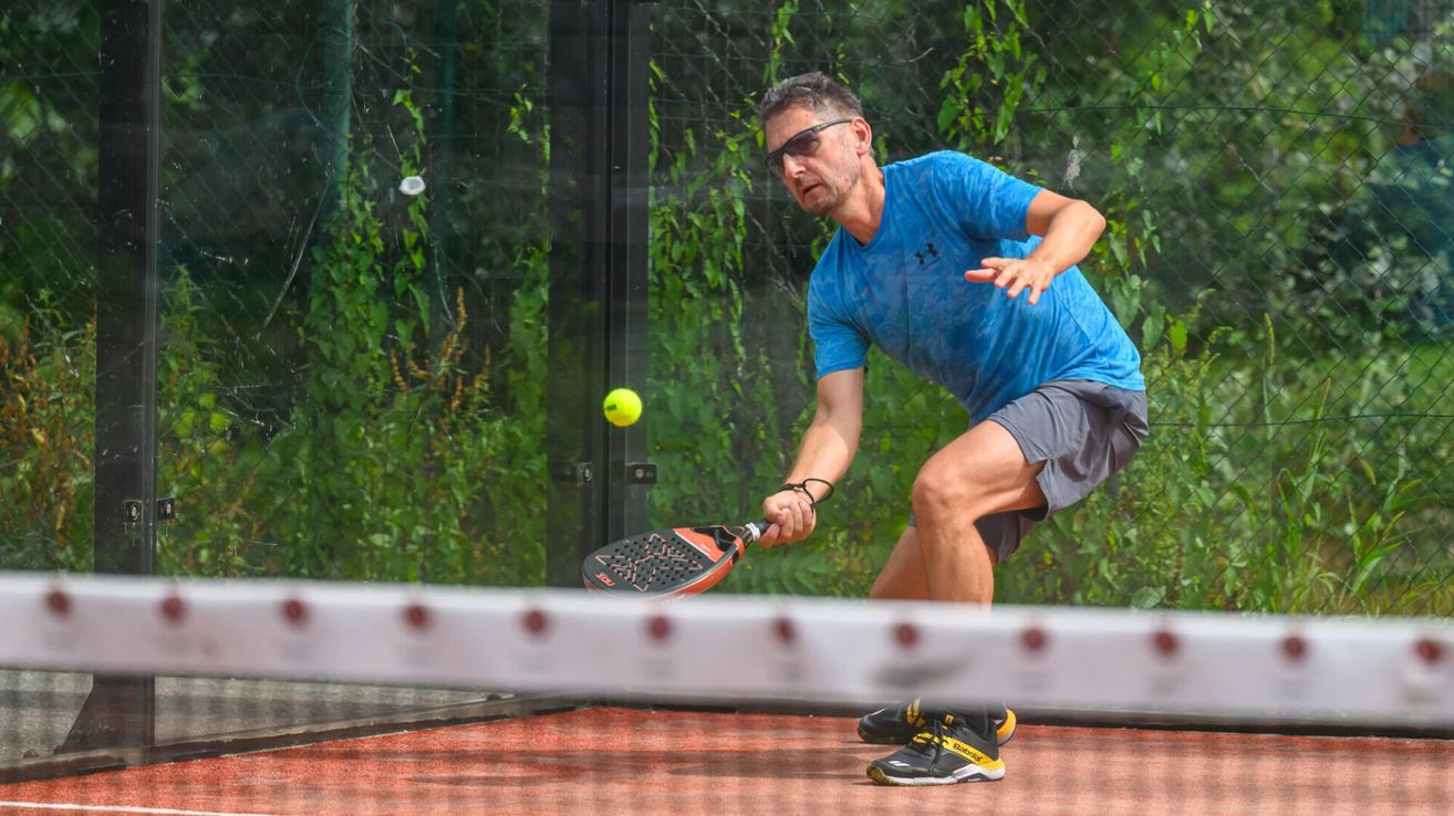 Ein Mann in einem blauen T-Shirt und grauen Shorts spielt Tennis auf einem Außenplatz und schlägt einen gelben Ball mit einem Schläger.