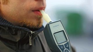 A man with a beard is blowing into an Alcoquant breathalyzer device. The device has a digital screen and buttons.