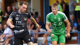 Zwei Fußballer im Einsatz, einer in Schwarz und der andere in Grün, mit einem verschwommenen Publikum im Hintergrund.