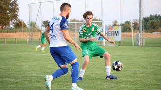 Zwei Fußballspieler sind auf einem Feld. Einer trägt ein blau-weißes Trikot und der andere ein grünes Trikot. Sie schauen beide auf den Ball.