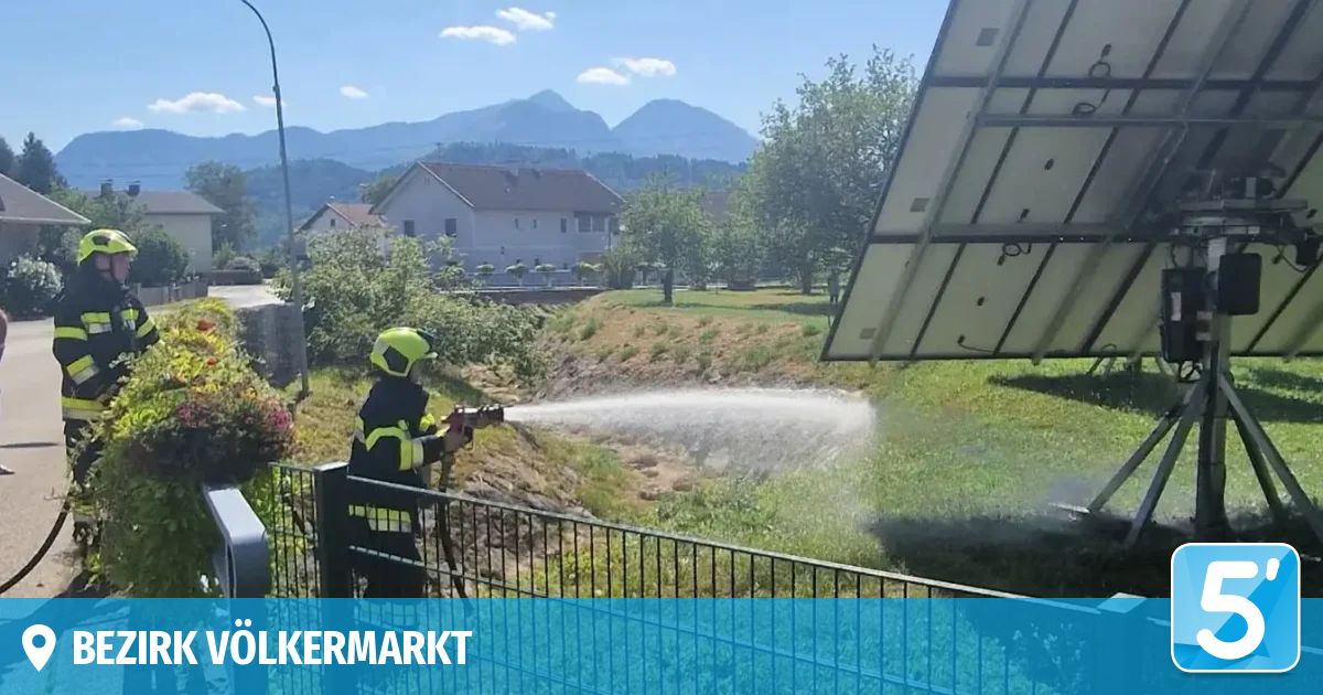 Ein Feuerwehrmann in voller Ausrüstung steht auf einem Zaun und spritzt Wasser aus einem Schlauch auf einen Wasserkörper. Im Hintergrund sind ein Haus und Solarpanele zu sehen.