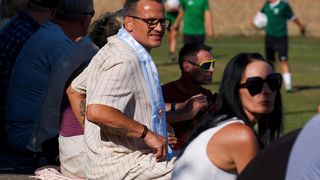 Ein Mann mit Brille und blauem Schal sitzt auf dem Rasen und lächelt. Hinter ihm schaut eine Frau in einem weißen Tanktop auf ihr Handy. Im Hintergrund stehen und spielen Leute Fußball.