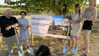 Drei Frauen halten Schaufeln vor einem Schild, das Hier Entsteht steht, mit einem schwarzen Hund im Vordergrund.