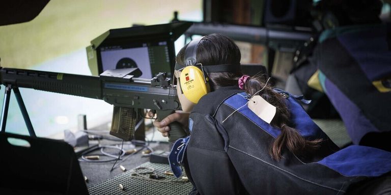 A woman is aiming a rifle at a target on a monitor, wearing ear protection and a blue jacket.