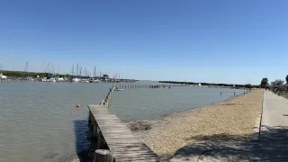 Eine Holzbrücke ragt in einen ruhigen Wasserkörper mit einem sandigen Strand und entfernten Booten. Der Himmel ist klar und blau.
