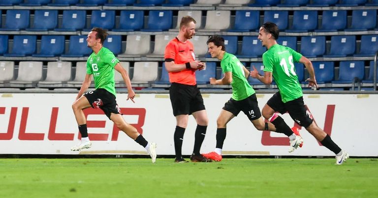 Vier Fußballspieler auf einem Feld, zwei in grünen Trikots laufen, einer in Orange pfeifend und einer in Grün blickt zurück. Leere blaue Tribünen im Hintergrund.