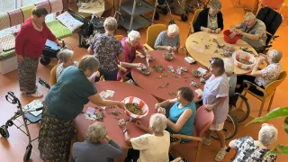 Eine Gruppe älterer Frauen sitzt um einen Tisch herum und ist mit einer Aktivität beschäftigt. Sie sitzen auf Stühlen und in Rollstühlen, mit Schüsseln voller Gegenstände und Scheren auf dem Tisch. Einige tragen Brillen und Halsketten.